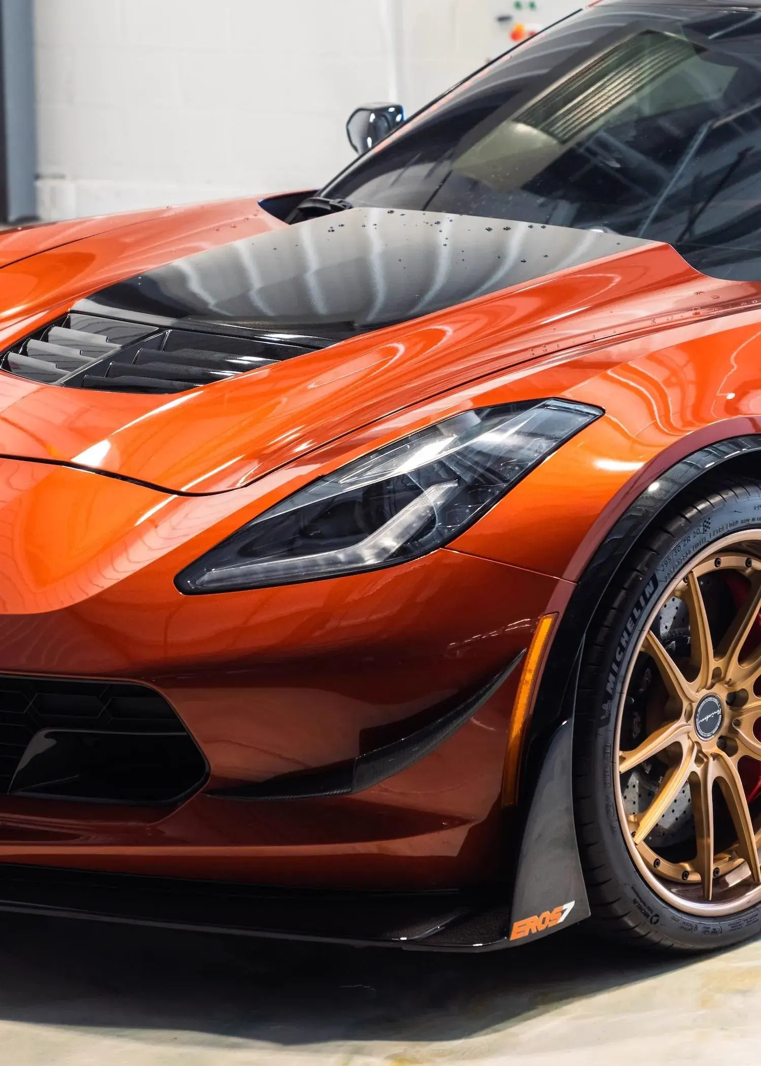 Close up of the front of an orange Corvette with a black hood insert and bronze wheels.
