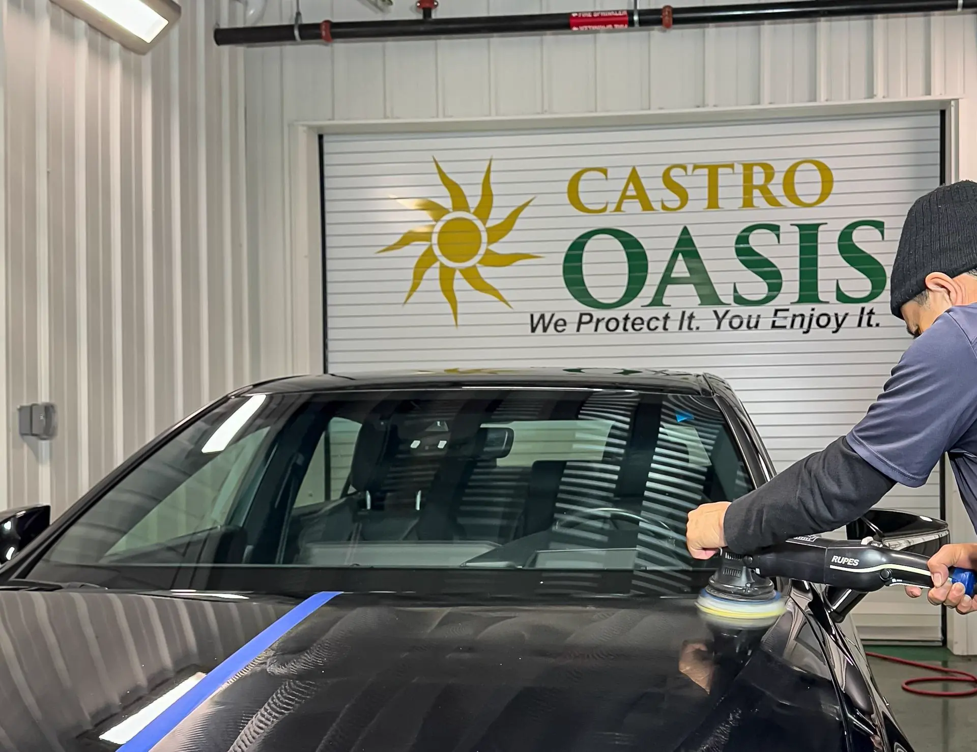 A technician machine polishing a dark hood in front of the shop's roller door, printed with the Castro Oasis sun logo and the line We Protect It. You Enjoy It.