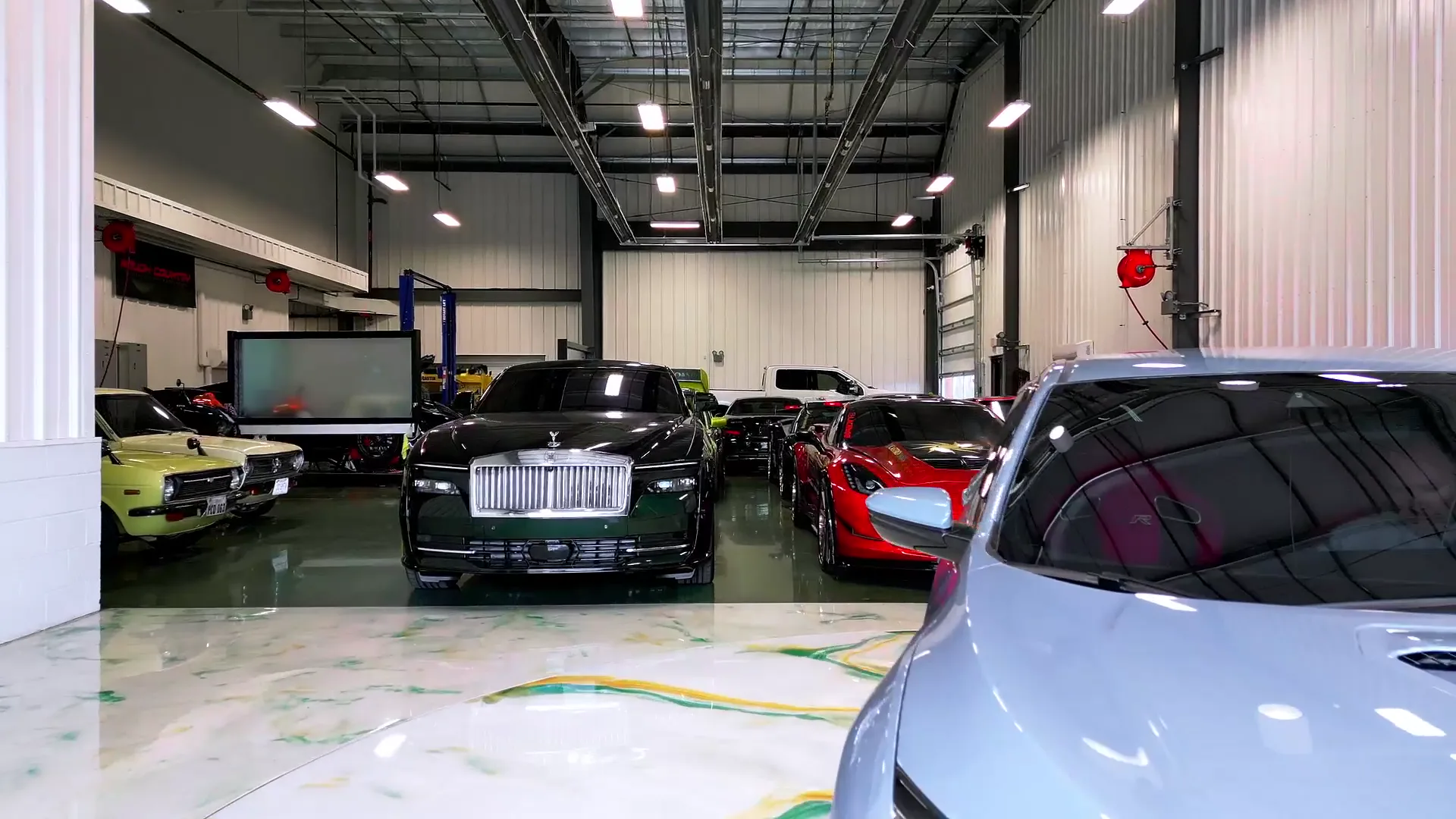 The showroom floor with a black Rolls-Royce, a green muscle car and a red supercar under a lit ceiling.