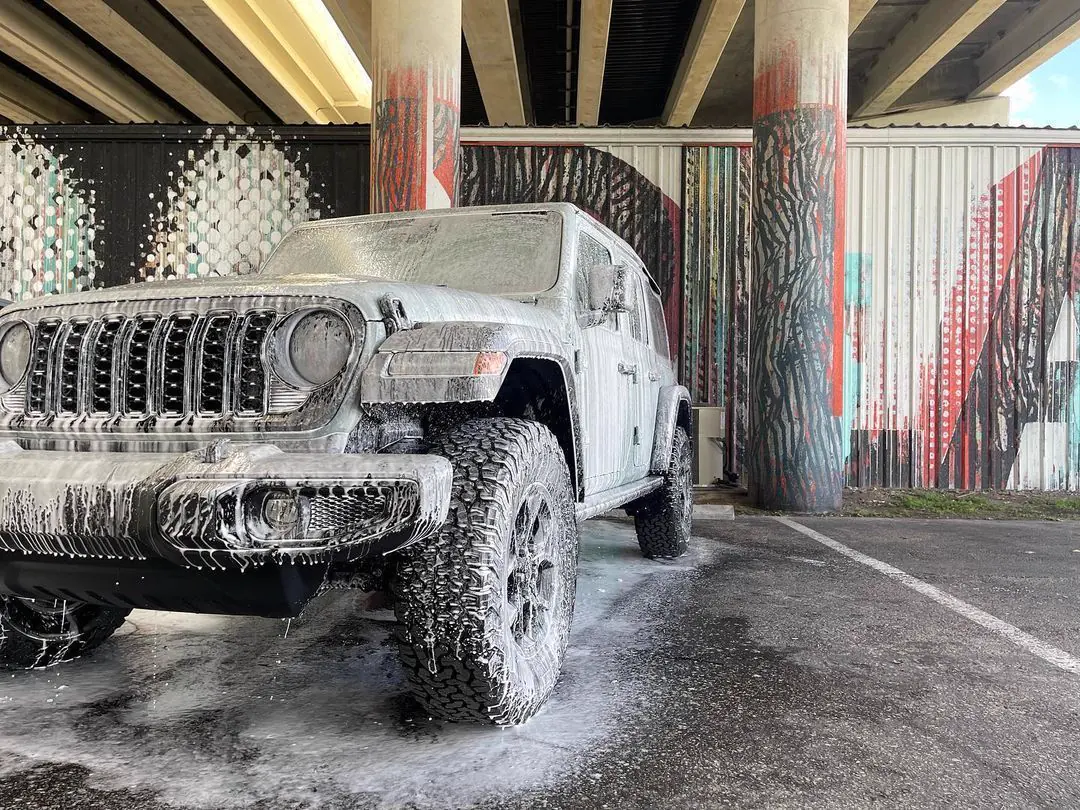 A white Jeep Wrangler covered in wash foam, parked under a highway overpass beside a painted mural.